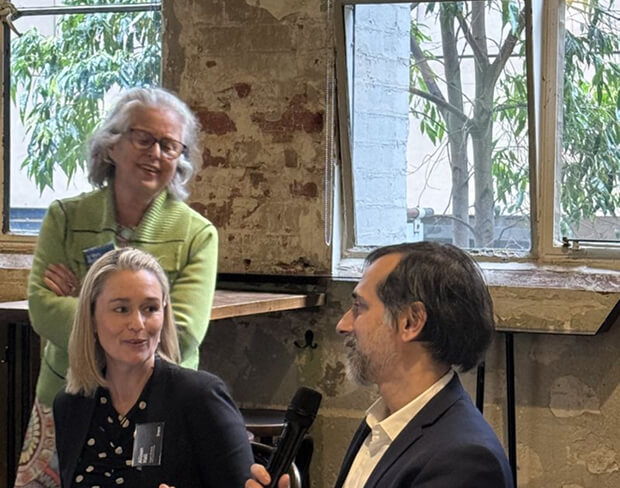 A man is seated, speaking into a microphone. Two woman smile on.